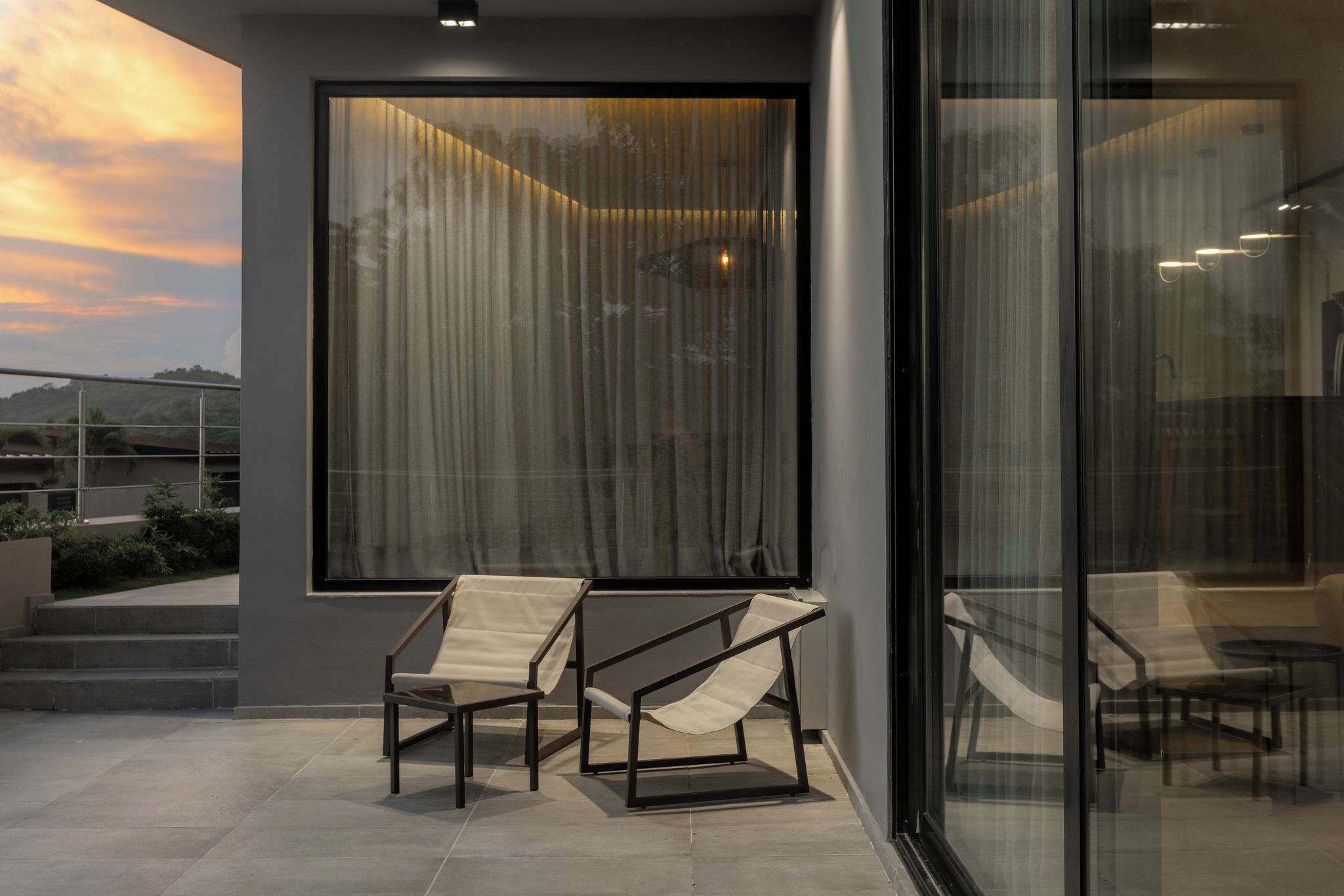 Un espace extérieur moderne aménagé avec deux chaises et une table, une grande fenêtre avec des rideaux et une vue sur le paysage.