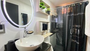 A modern bathroom with a round mirror, white sink, black countertop, and a glass shower enclosure. - Comfortable apartment and excellent location (Barranquilla)