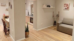 Ett modernt vardagsrum med en soffa, en matplats med stolar, ett kök med diskho och skåp samt en hall som leder till ett annat rum.