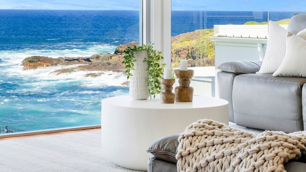 Un salon moderne avec vue sur l’océan, comprenant un canapé gris, une petite table d’appoint ronde et blanche agrémentée d’objets décoratifs, et un plaid tricoté.
