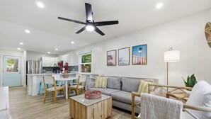 Ein modernes Wohnzimmer mit Sofa, Couchtisch und Sesseln, ein Essbereich mit Tisch und Stühlen, eine Küche mit weißen Schränken und einem Deckenventilator.