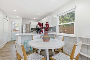 A modern kitchen with white cabinetry, a stainless steel refrigerator, and a vase with red flowers on a round table. - Toes in the Sand Retreat II (Jacksonville Beach)