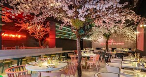 Ein Restaurant mit rosafarbenen Stühlen, runden Tischen und einem Baum mit blühenden rosa Blüten.