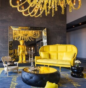 Ein luxuriöses Zimmer mit einem großen Spiegel, der eine goldene Statue, ein gelbes Sofa, einen schwarzen Hocker und einen Chandelier reflektiert.