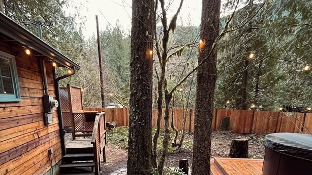 Une cabane en bois dotée d’un patio en pierre, d’un bain à remous et d’un espace salon couvert, le tout entouré d’arbres.