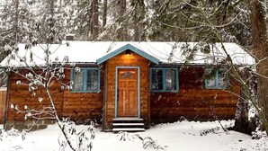 Une cabane en bois, dont le toit est recouvert de neige, entourée de grands arbres à feuilles persistantes.