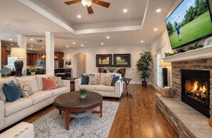 A living room with a sofa, coffee table, fireplace, TV, and kitchen in the background. - 4 Ensuites, Championship Golf, Pool, Hottub, 30ft Patio with Outdoor Dining. (Branson)