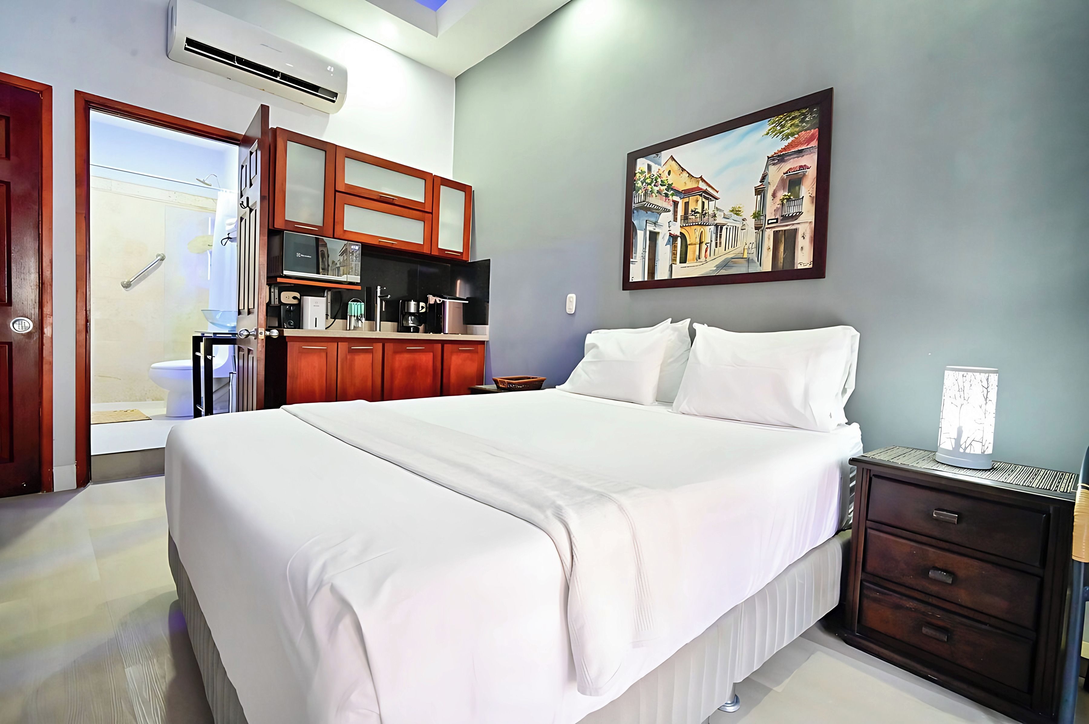A hotel room with a bed, a nightstand, a painting, a kitchenette, and a bathroom.
