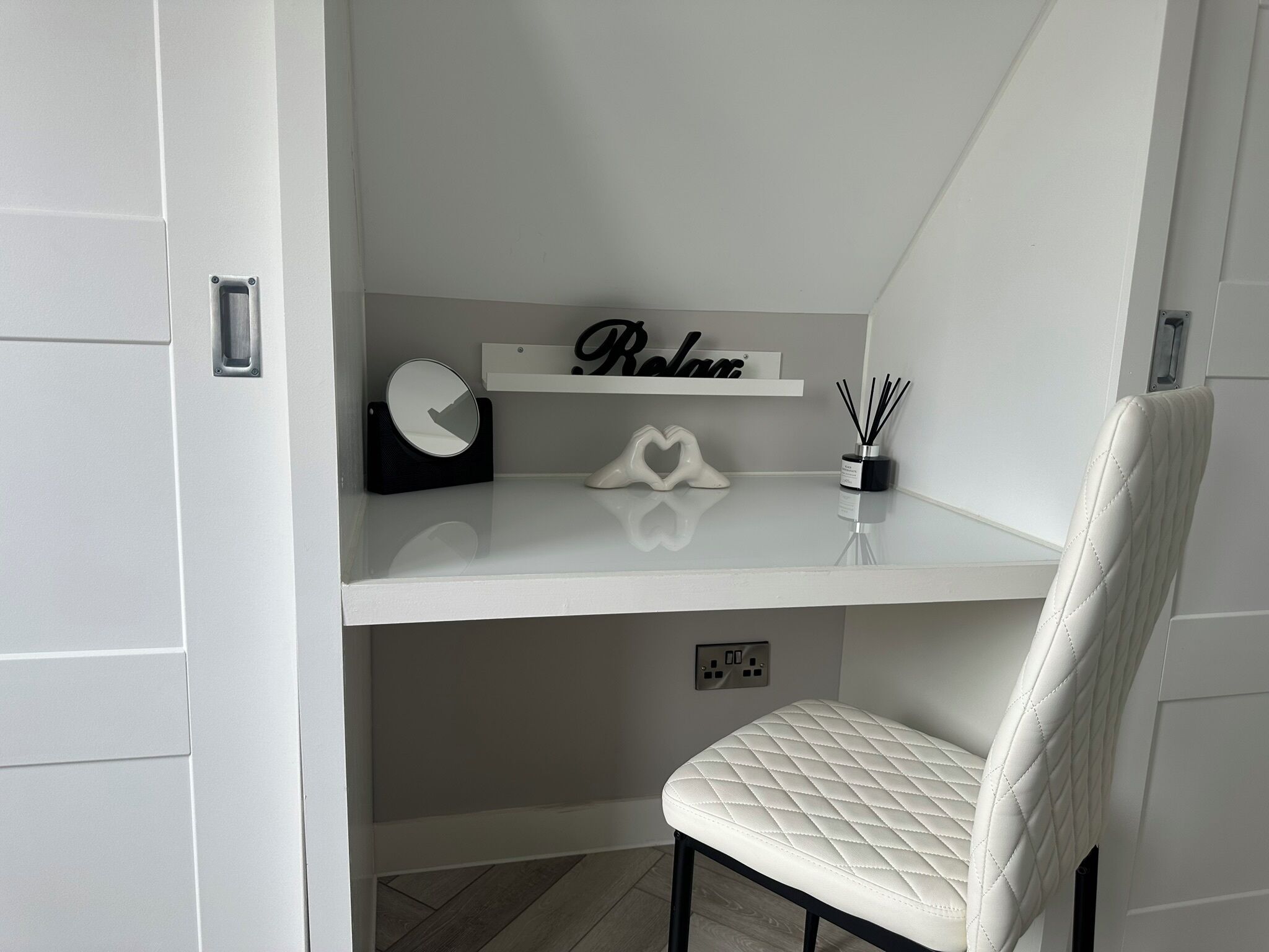 Una habitación moderna y luminosa con una silla blanca, un estante con un reloj y una decoración con forma de corazón, y un pequeño estante con un letrero y difusores de aroma.