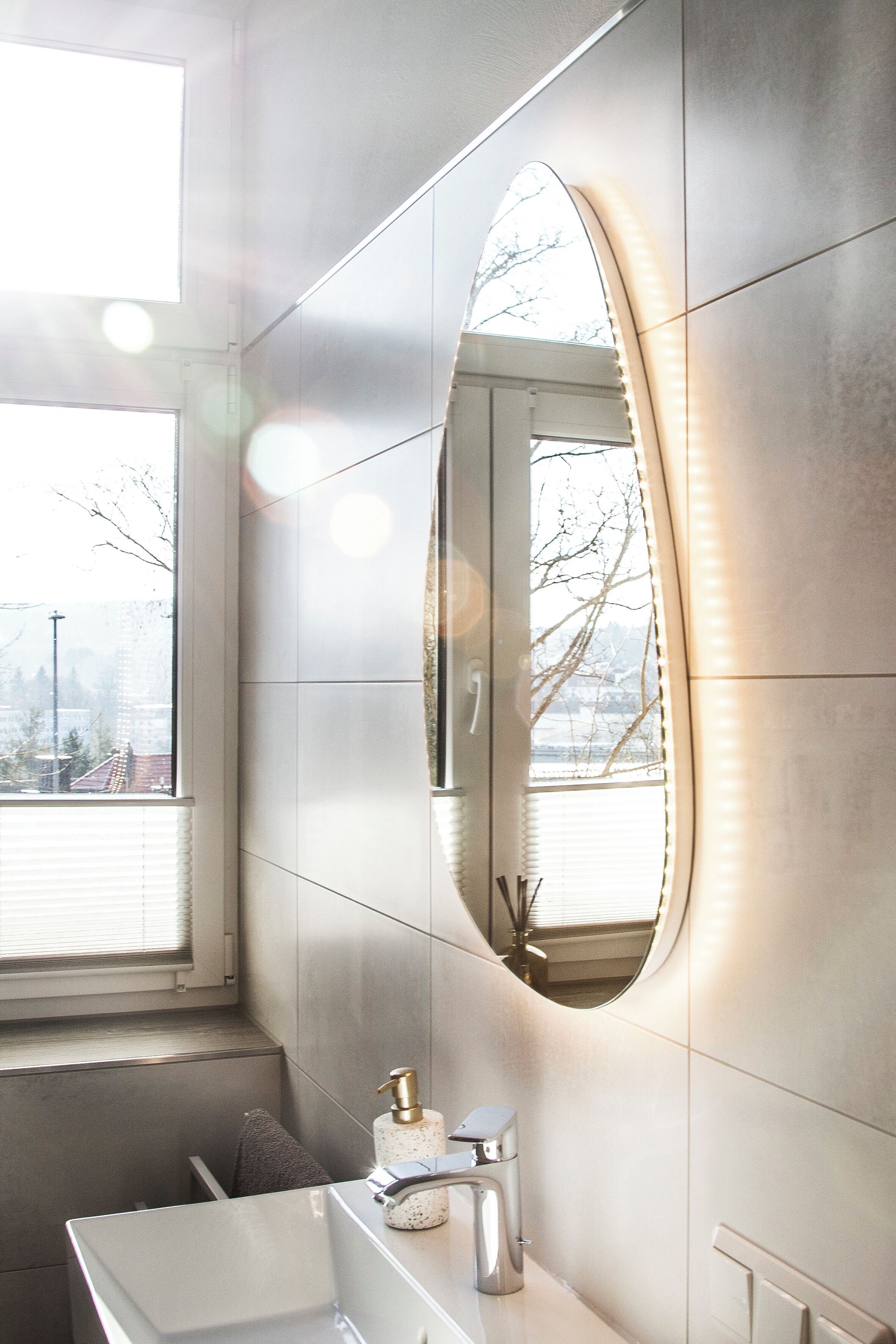 Ein modernes Badezimmer mit einem großen Spiegel, einem weißen Waschbecken und einem Fenster mit Jalousien.