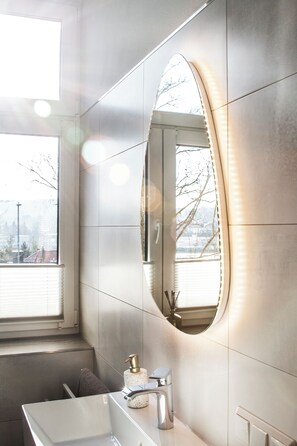 A modern bathroom with a large mirror, a white sink, and a window with blinds. - Vacation apartment in Suhl - Wilhelminian style villa (Suhl)