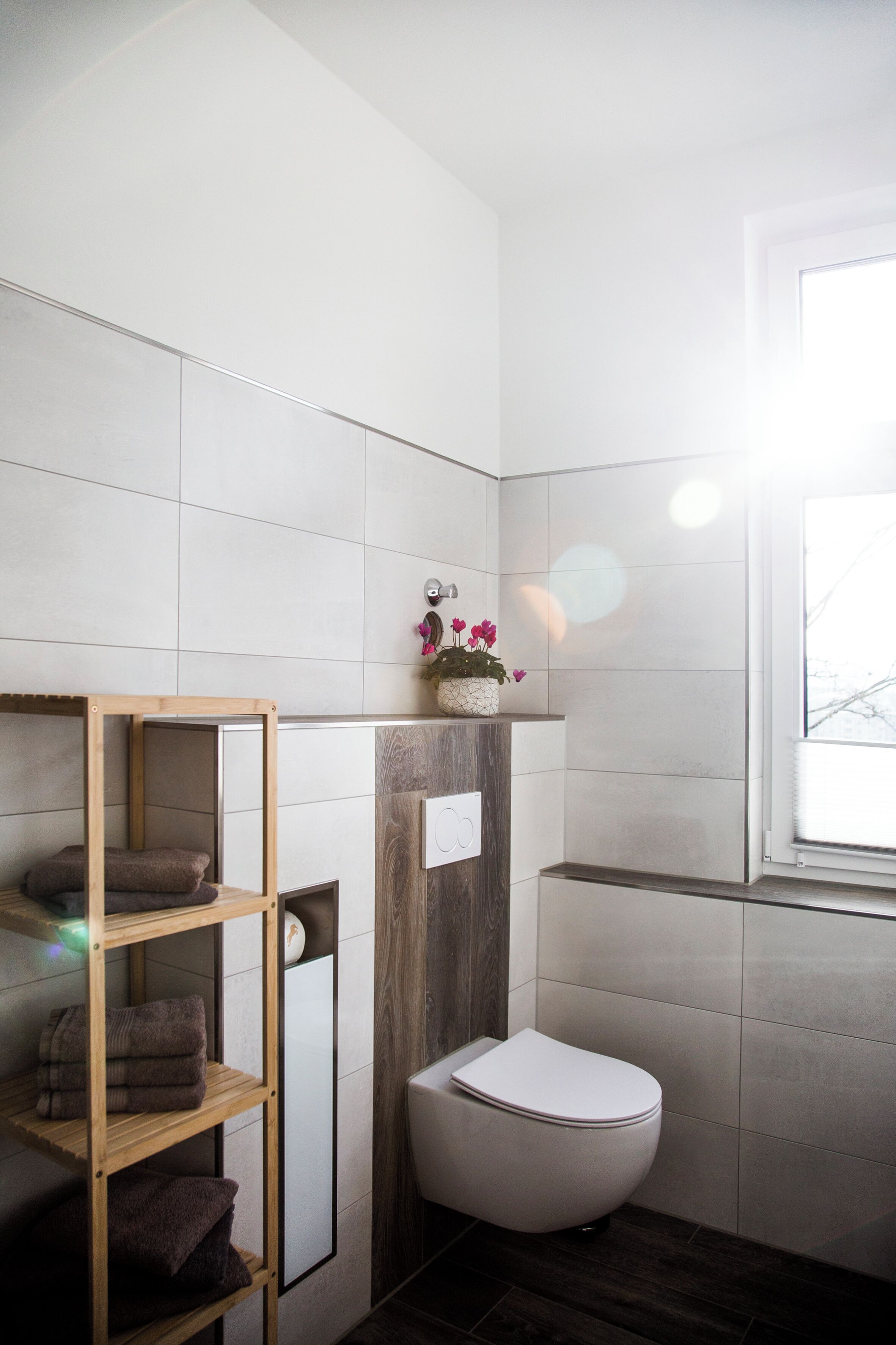 A modern bathroom with white tiles, a wooden shelf with towels, a white toilet, and a window with natural light.