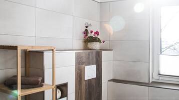 A modern bathroom with white tiles, a wooden shelf with towels, a white toilet, and a window with natural light.