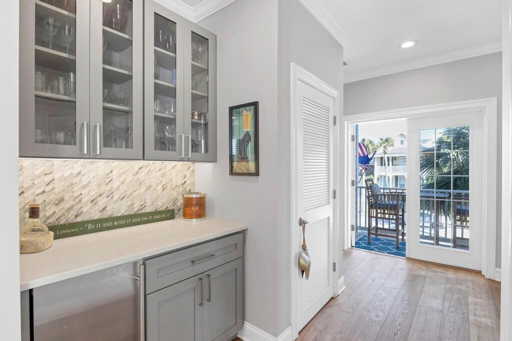 A kitchen with grey cabinets, a marble countertop, and a door leading to a balcony with a view of palm trees and houses.