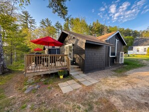 A cabin with a wooden deck, a covered patio with red umbrellas, and a gravel driveway. - Riverside Cottage • Private Beach • Mountain Views (Bartlett)