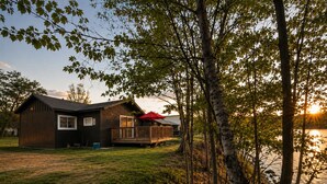 A cabin with a deck, a large window, and a view of a lake at sunset. - Riverside Cottage • Private Beach • Mountain Views (Bartlett)