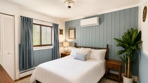 A bedroom with a bed, bedside tables, a lamp, an air conditioner, a window with curtains, a potted plant, and a wooden dresser. - Riverside Cottage • Private Beach • Mountain Views (Bartlett)