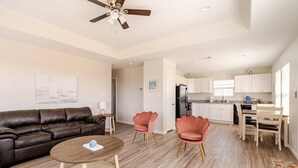 A modern living room with a brown leather sofa, a round wooden coffee table, and two pink chairs. A ceiling fan is visible above. - Sunset Bellini - Hot Tub, Golf Cart, Dog Friendly! (Bolivar Peninsula)