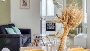 A modern living room with a sofa, a small round table, and a vase with dried plants. A kitchen with white cabinets and a spiral staircase is visible in the background. - Louis, maison de ville en copro (Rouen)