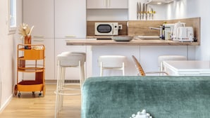 A modern kitchen with white cabinets, a wooden countertop, and a built-in oven. There are two white bar stools and a green sofa in the foreground. - Les Mathurins, F2 unique terrasse, parking et sport (Rouen)
