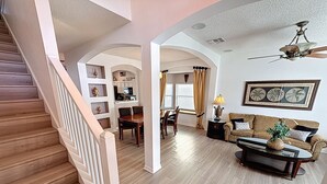 A spacious living area with a wooden staircase, a dining table with chairs, a sofa, and a ceiling fan. - Tinkerbells Suites, 4m Disney, 6/5.5 Large pool+Spa, 4 themed masters, pets (Kissimmee)