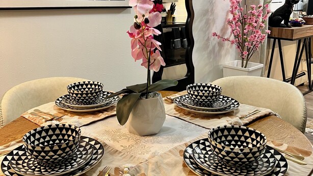 A dining table set with black and white patterned plates, a vase with pink flowers, and a mirror on the wall.