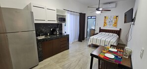 Ein kompaktes Hotelzimmer mit Küchenzeile, einem Bett mit gestreifter Bettwäsche, einem Deckenventilator und einem an der Wand montierten Fernseher.