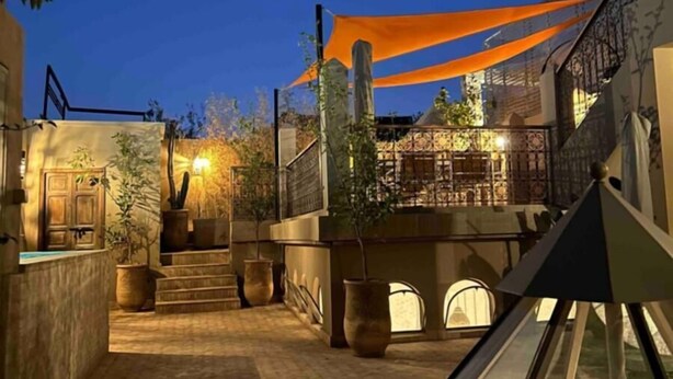 Ein Innenhof mit einer Treppe, die zu einem Balkon führt, umgeben von Blumentöpfen und einem großen, orangefarbenen Sonnensegel.
