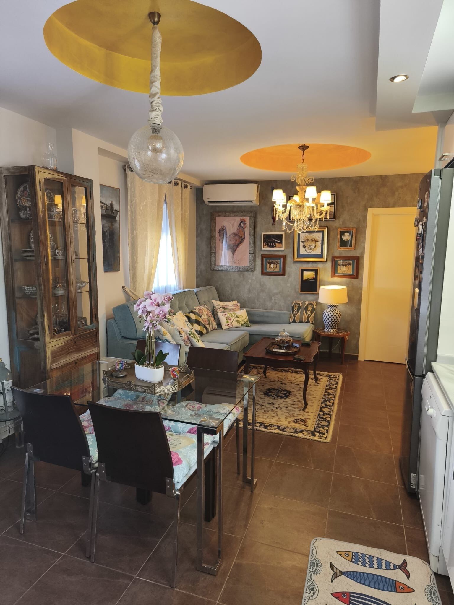Un salon comprenant un canapé, une table basse, une table à manger avec des chaises, un lustre et un réfrigérateur.