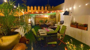 Un espace extérieur aménagé pour prendre les repas, avec une table dressée pour deux personnes, entourée de chaises. On y trouve des plantes en pot et des guirlandes lumineuses.