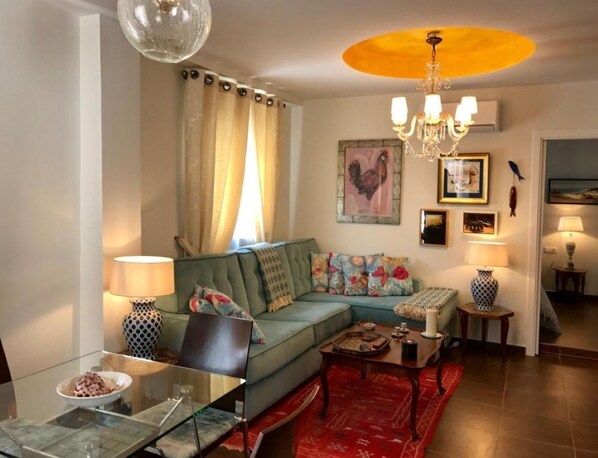 Un salon chaleureux avec un canapé vert, une table basse en verre et un tapis rouge. On y trouve un lustre et un tableau représentant un coq accroché au mur.