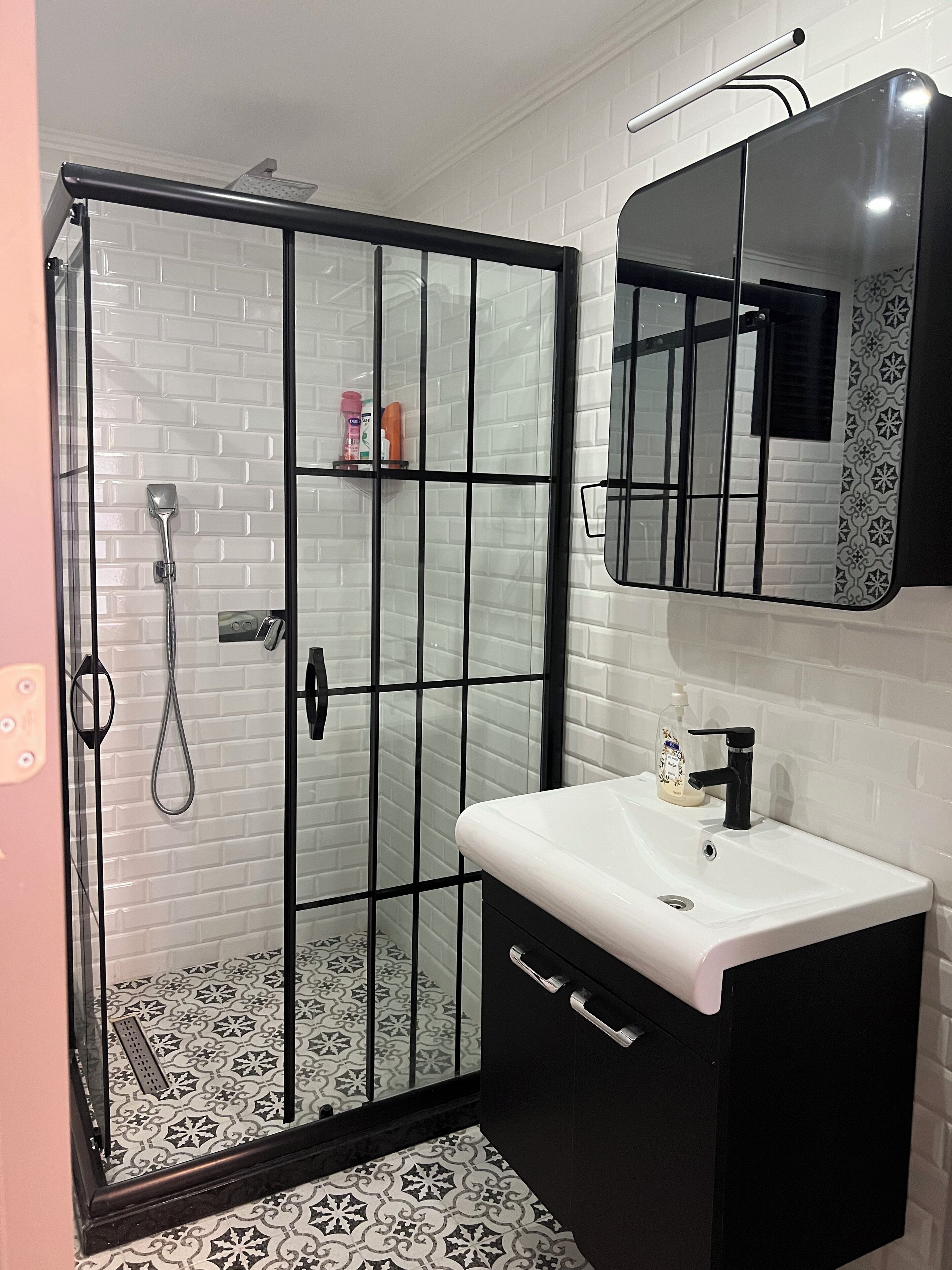 Un baño con ducha de vidrio, lavamanos blanco con grifo negro y espejo con marco decorativo.