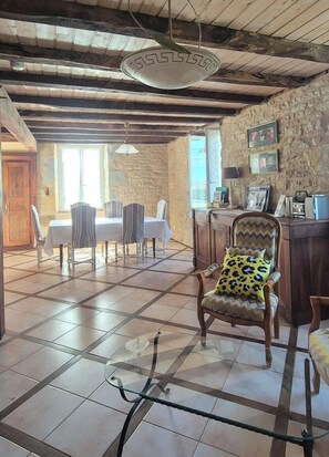 Un coin repas rustique avec un plafond en bois, des murs en pierre et un sol carrelé. On y trouve une table en verre avec des chaises, une chaise en bois avec un coussin à motifs léopard, et un mur en pierre orné de tableaux encadrés.