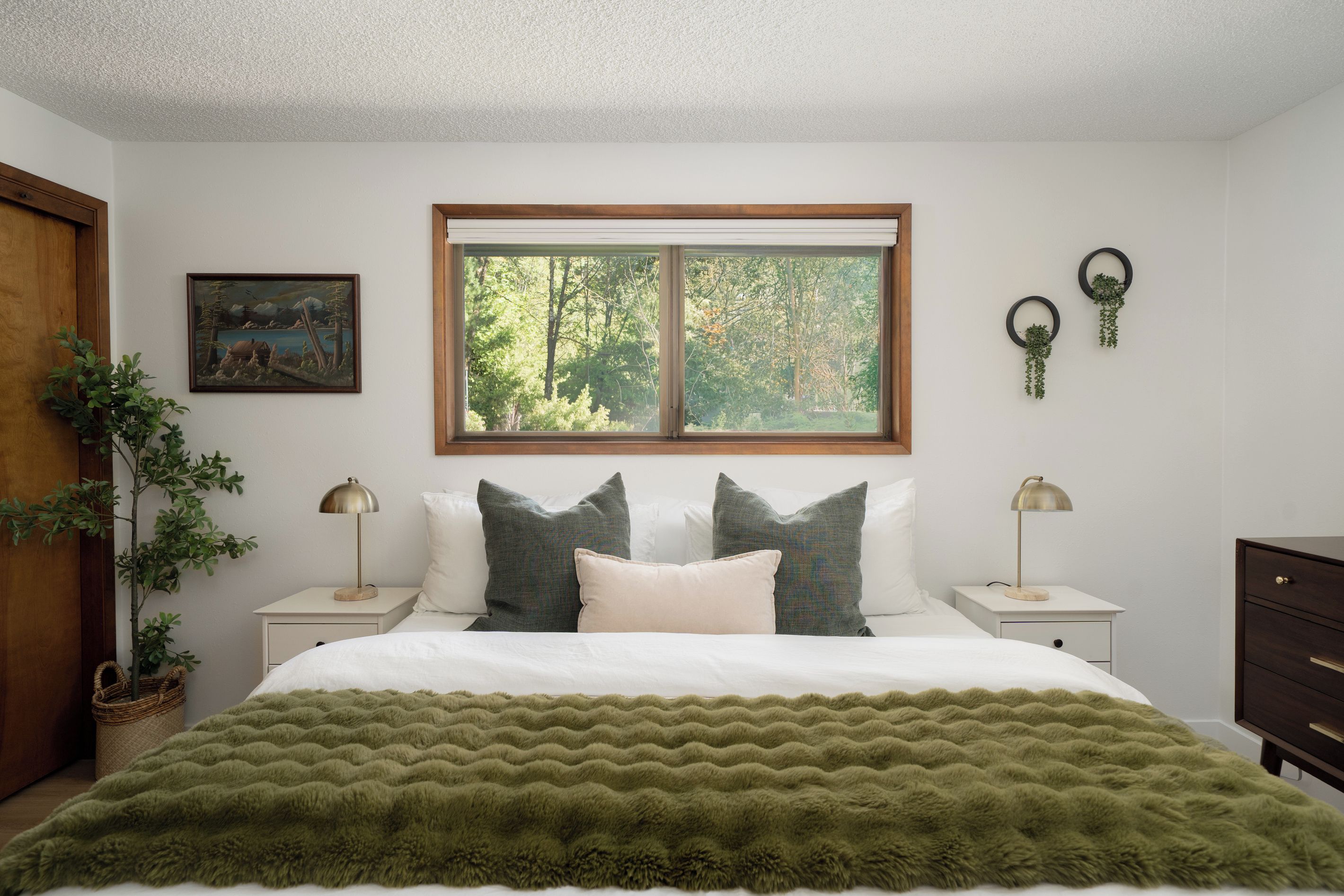 Une chambre à coucher avec un lit, deux tables de chevet, une fenêtre, un tableau et une plante.