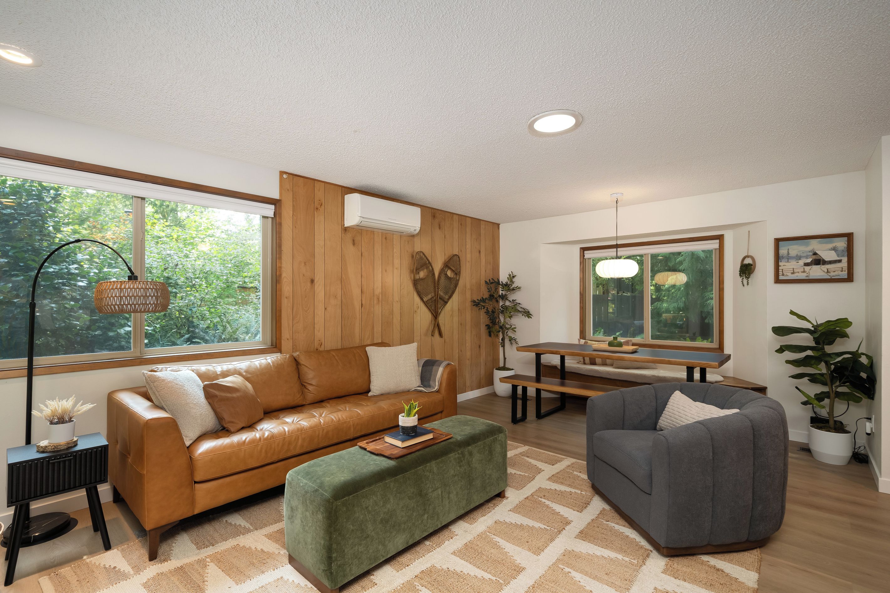 A modern living room with a leather sofa, a green ottoman, a grey armchair, a wooden dining table, and a large window with a view of greenery.