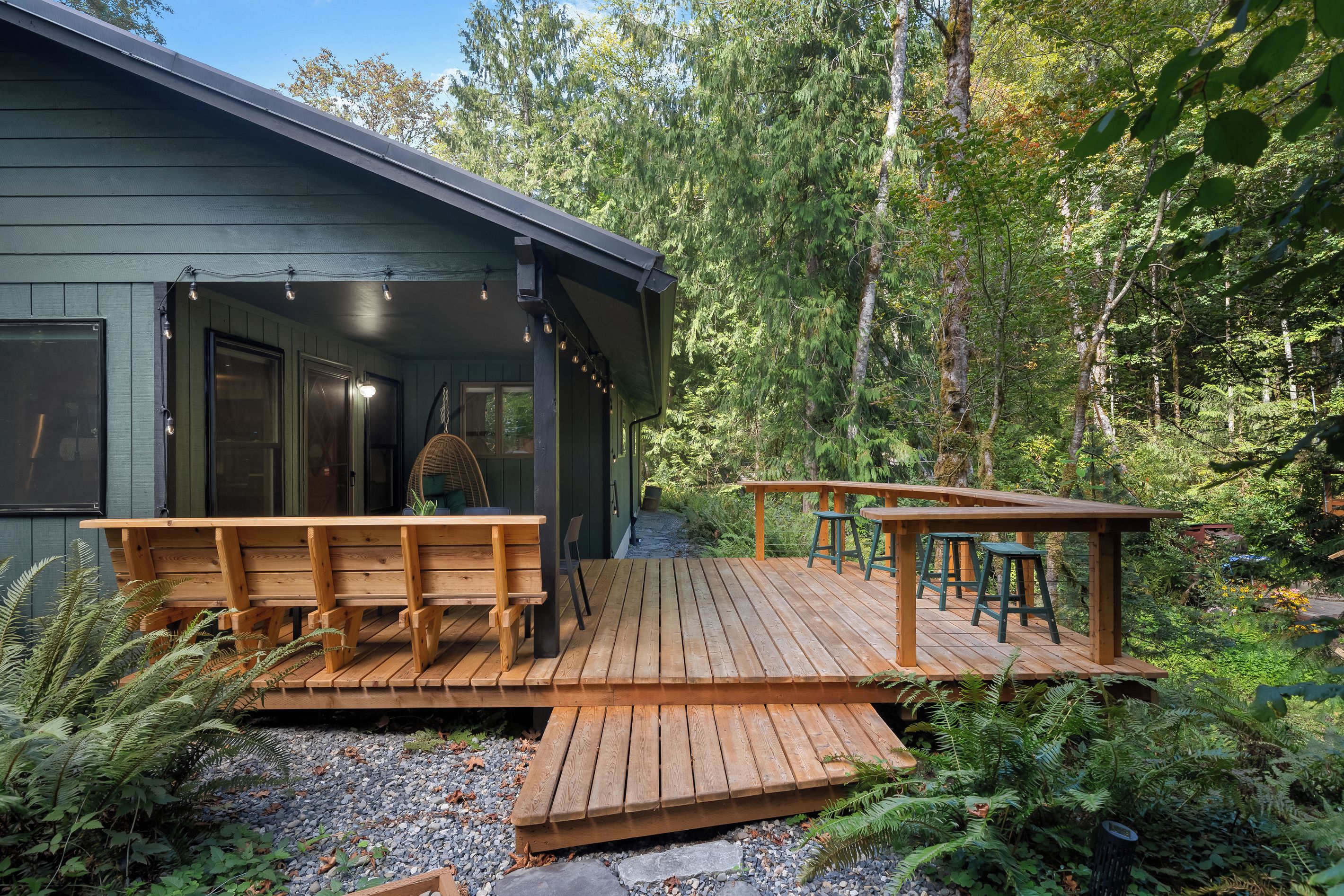 A wooden deck with a bench and stools, surrounded by dense forest.