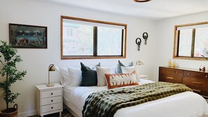 Una camera da letto con un letto, comodini, una cassettiera, una finestra con persiane, una luce a soffitto, una sveglia a muro e un quadro incorniciato.