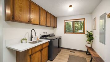 A modern kitchen with wooden cabinets, a stainless steel sink, a built-in washing machine, and a window with a view of greenery.