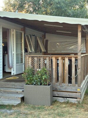 A covered outdoor area with a wooden deck, a large planter with plants, and a view of the house in the background.