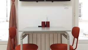 Een kamer met een aan de muur bevestigde televisie, een tafel met twee rode stoelen en een raam met gordijnen.