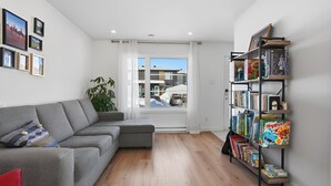 Ein modernes Wohnzimmer mit einem grauen Ecksofa, einem Bücherregal voller Brettspiele und einem großen Fenster mit Blick auf Gebäude.