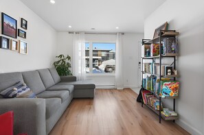 Ein modernes Wohnzimmer mit einem grauen Ecksofa, einem Bücherregal voller Brettspiele und einem großen Fenster mit Blick auf Gebäude.