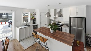 Eine moderne Küche mit weißen Schränken, einer Holz Arbeitsplatte und Edelstahlgeräten. Zwei Pendelleuchten, ein Fenster mit Blick ins Freie und ein Topf mit Pflanze auf der Arbeitsplatte sind vorhanden.