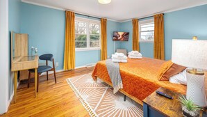 A hotel room with a bed, a desk, a chair, a TV, and a window with curtains. - Stylish Downtown Bungalow with Fire Pit (Carrboro)