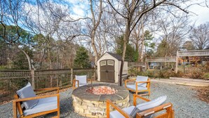 A stone fire pit surrounded by wooden chairs with cushions, set on gravel. - Stylish Downtown Bungalow with Fire Pit (Carrboro)