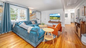 A modern living room with a blue sofa, a small round table, and an orange chair. There is a TV mounted on the wall and a window with curtains. - Stylish Downtown Bungalow with Fire Pit (Carrboro)