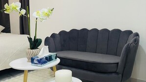 Et moderne hotelværelse med en grå sofa, et lille rundt bord med et stearinlys og en vase med hvide blomster.