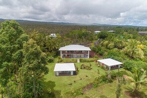 Ein modernes, zweistöckiges Haus mit einem großen Balkon, umgeben von üppiger Vegetation und einer hügeligen Landschaft.