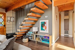 A modern interior with a wooden staircase, a couch, a bookshelf, and a framed artwork. - 2BR Chic A-Frame w Slopes Views near Bear Mountain (Big Bear Lake)