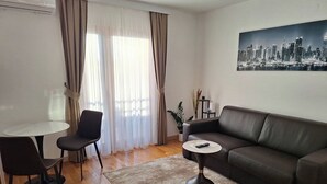 Ein modernes Wohnzimmer mit einem braunen Ledersofa, einem kleinen runden Couchtisch und einem weißen Tisch mit zwei Stühlen. Ein großes Fenster mit transparenten Vorhängen lässt viel natürliches Licht herein.